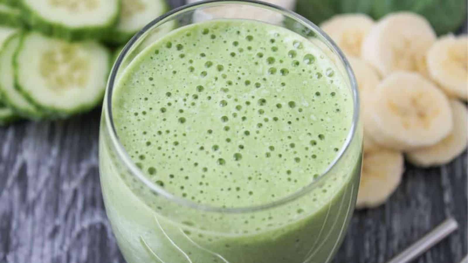 A glass of green smoothie with a frothy top, placed on a wooden surface, surrounded by sliced cucumber and banana.