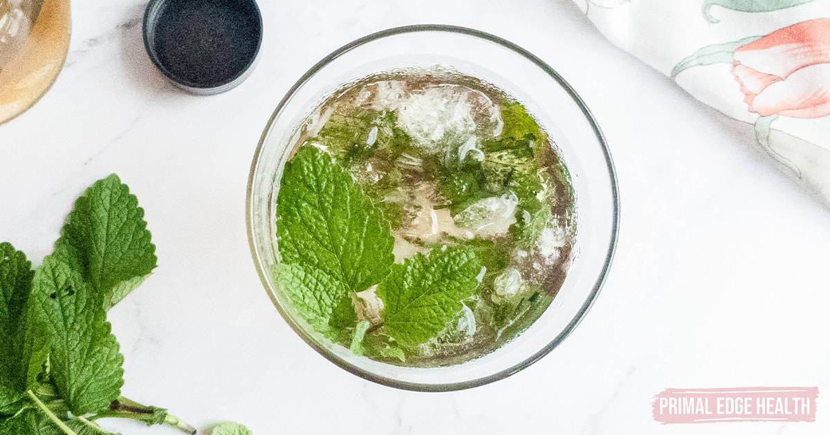 Top-down view of a glass filled with a clear iced beverage garnished with fresh mint leaves, placed on a white surface next to a sprig of mint and a floral cloth.
