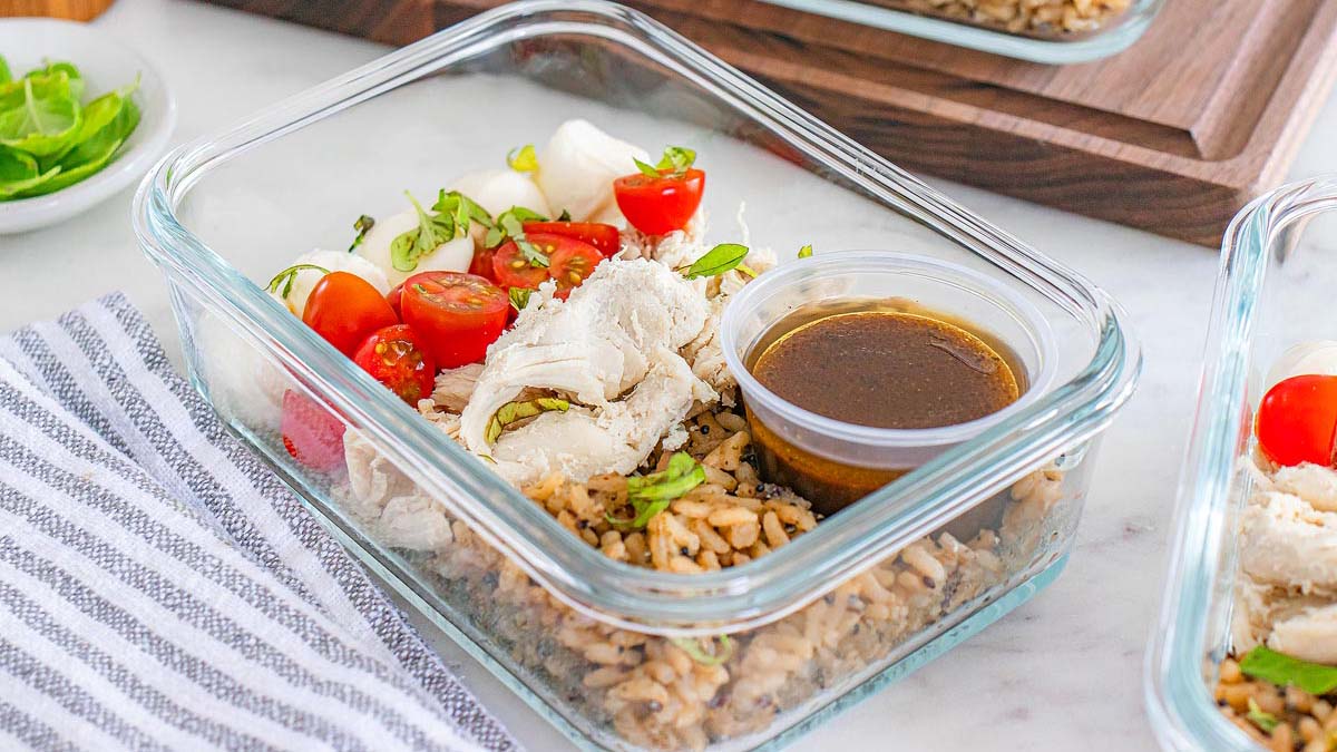 A glass meal prep container with sliced chicken, cherry tomatoes, mozzarella, brown rice, and a small container of dark dressing on a striped cloth.