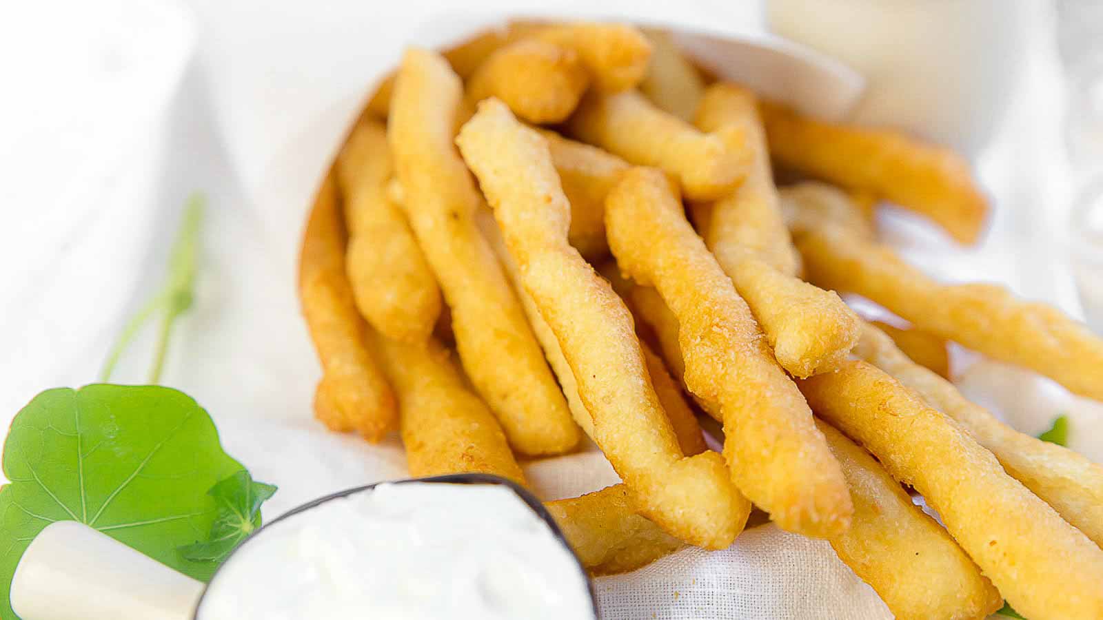 A pile of golden, crispy fries is served on a white surface with a side of white dipping sauce and a green leaf garnish.