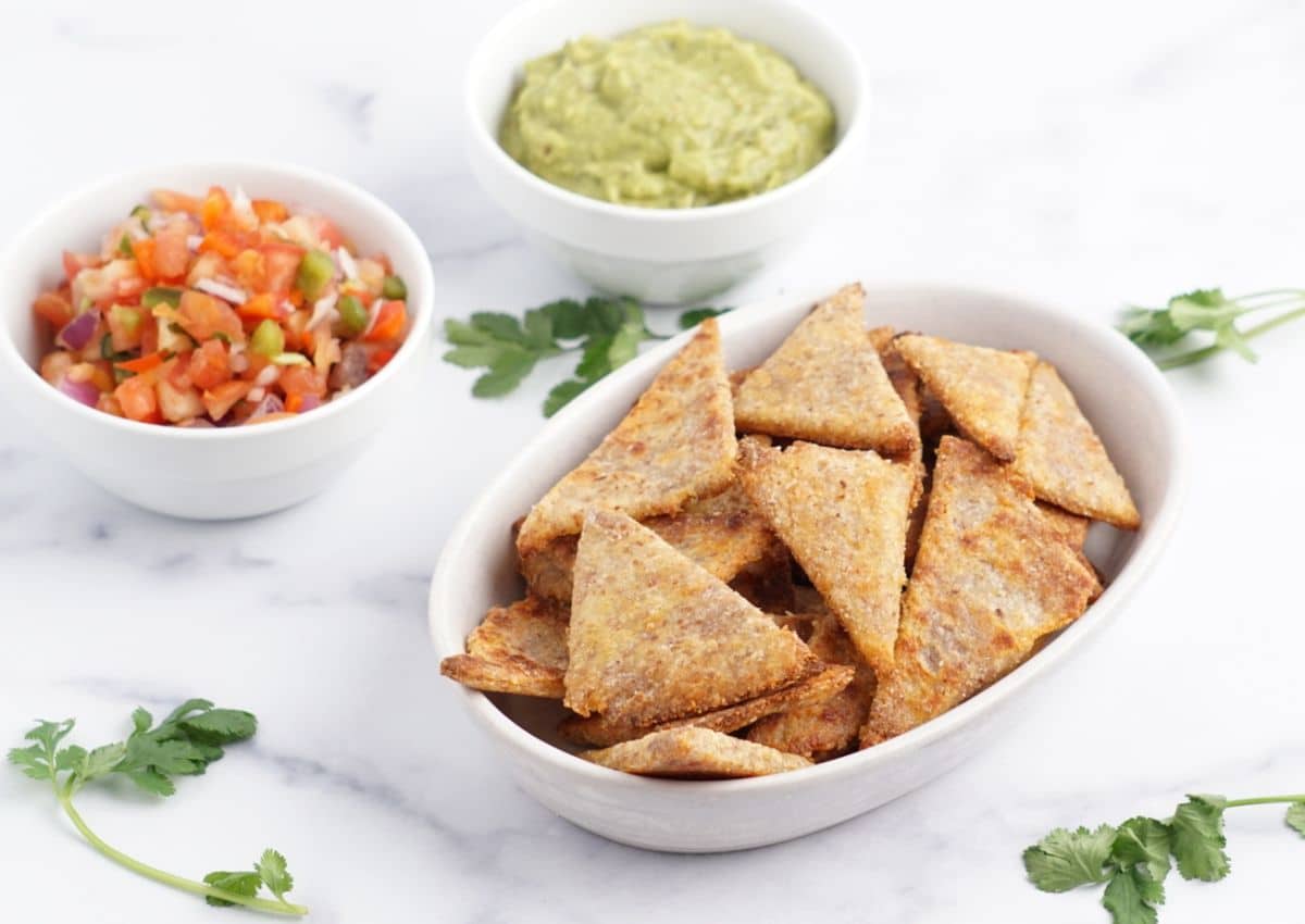 A white dish of triangular tortilla chips with bowls of guacamole and salsa on a marble surface, garnished with cilantro leaves.