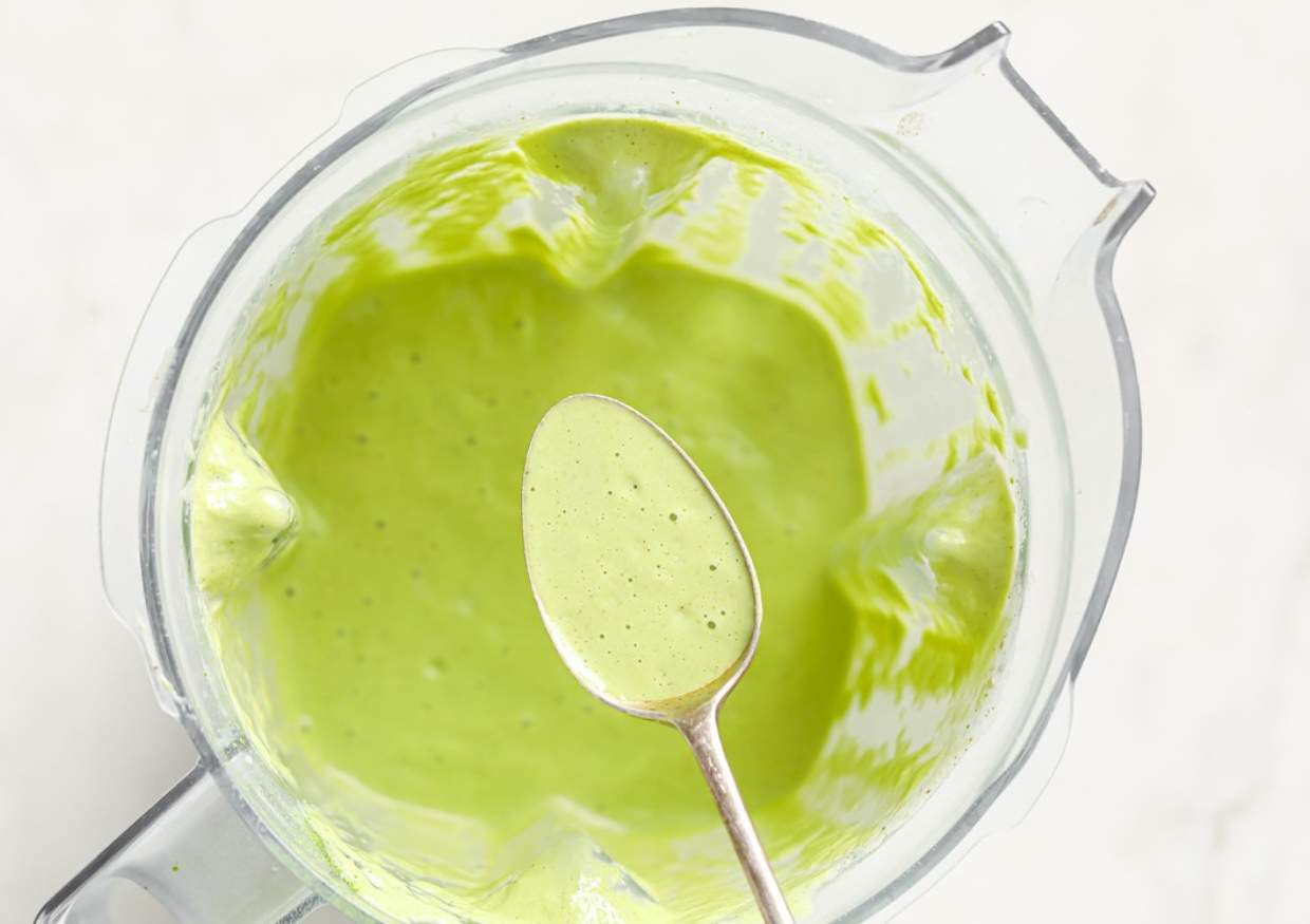 A metal spoon rests above a blender containing a smooth, light green sauce or smoothie.