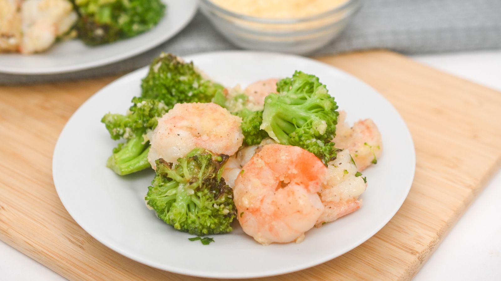 A white plate with cooked shrimp and broccoli sits on a wooden surface, with a bowl of grated cheese and another plate in the background.