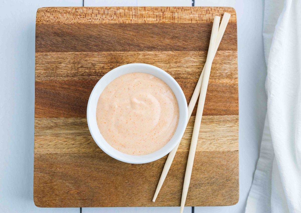 A small white bowl filled with creamy orange sauce sits on a wooden board, with a pair of chopsticks placed beside it.