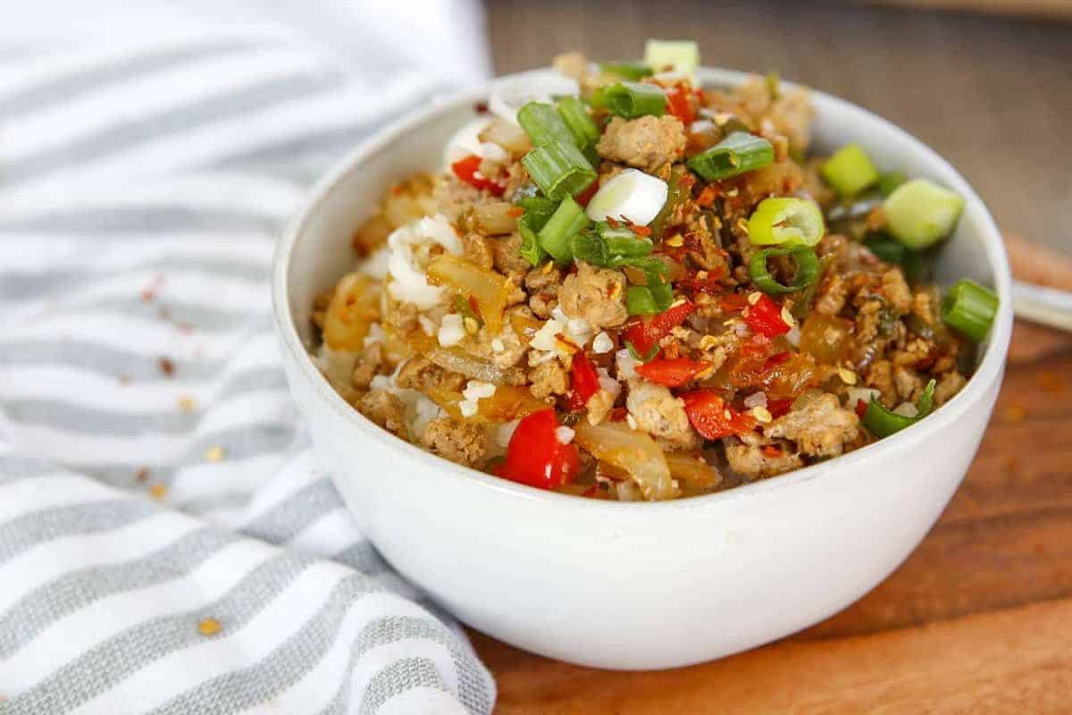 A white bowl filled with a stir-fry dish featuring ground meat, diced red peppers, green onions, and seasonings, set on a wooden surface next to a striped cloth.