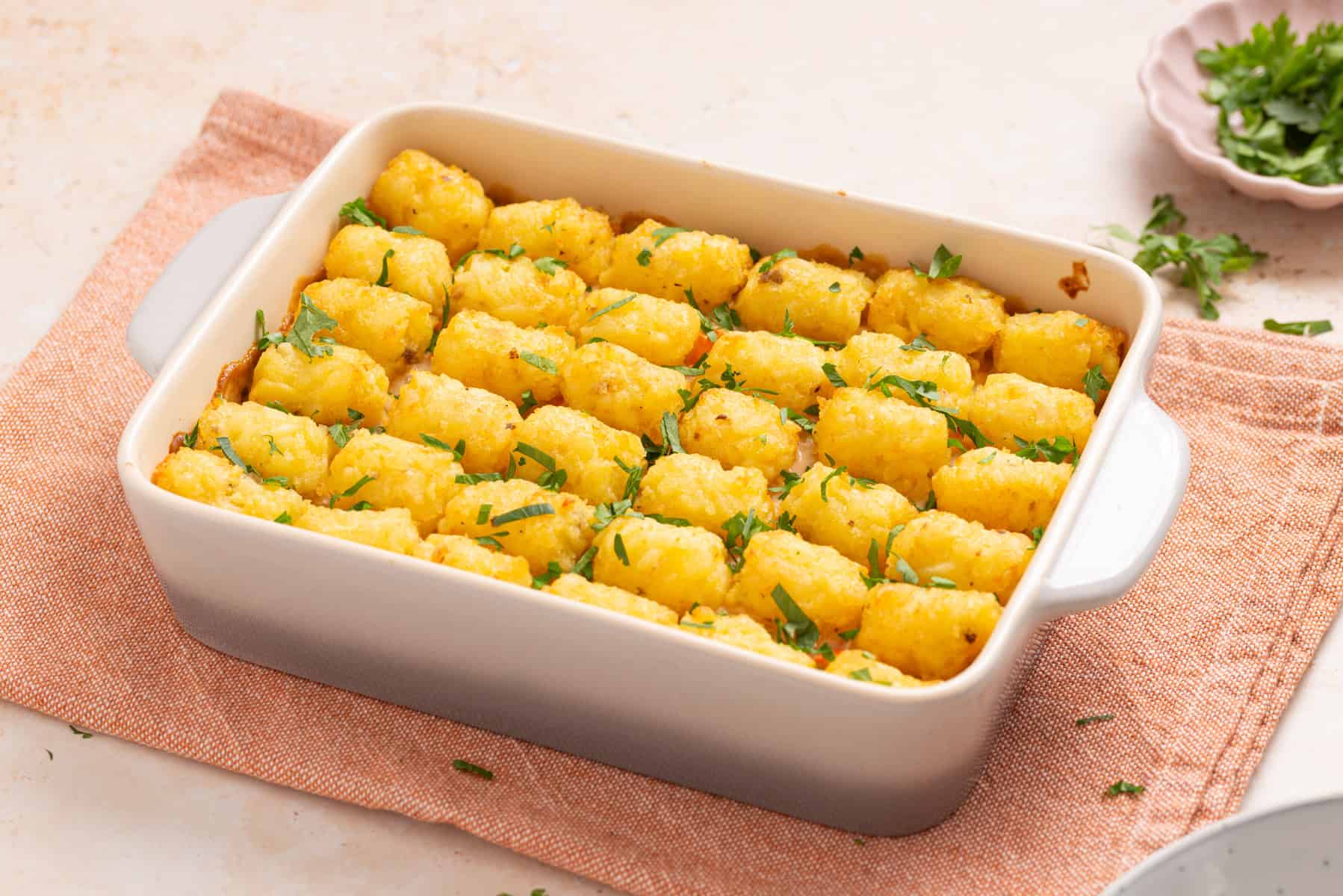 A white baking dish filled with tater tot casserole, topped with evenly arranged tater tots and garnished with chopped parsley, sits on a pink cloth.