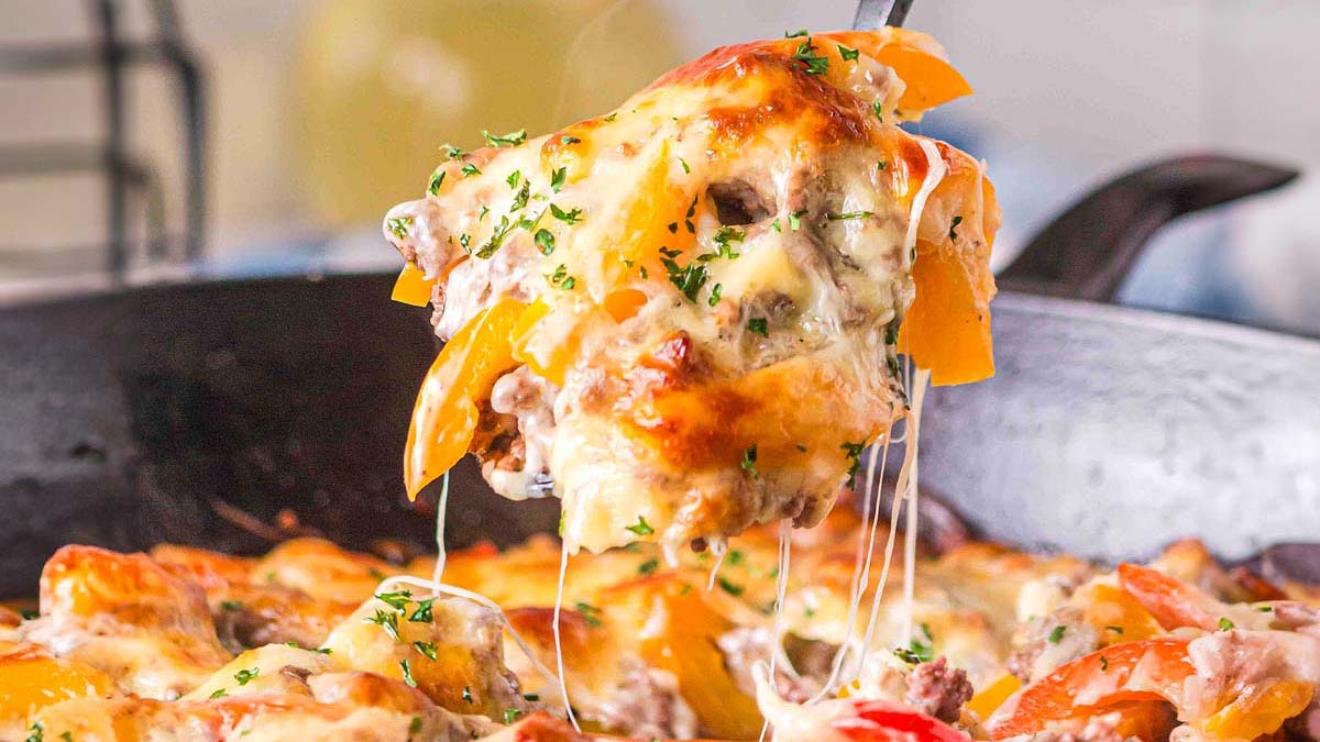 A spoon lifts a cheesy baked dish with melted cheese, ground meat, and sliced bell peppers from a skillet.