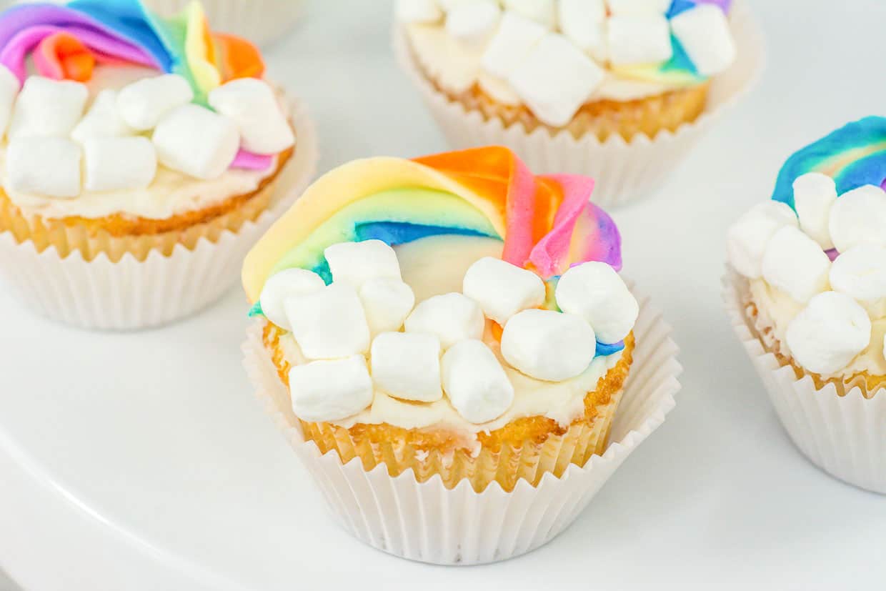 Cupcakes topped with white frosting, mini marshmallows, and rainbow-colored icing, arranged on a white surface.