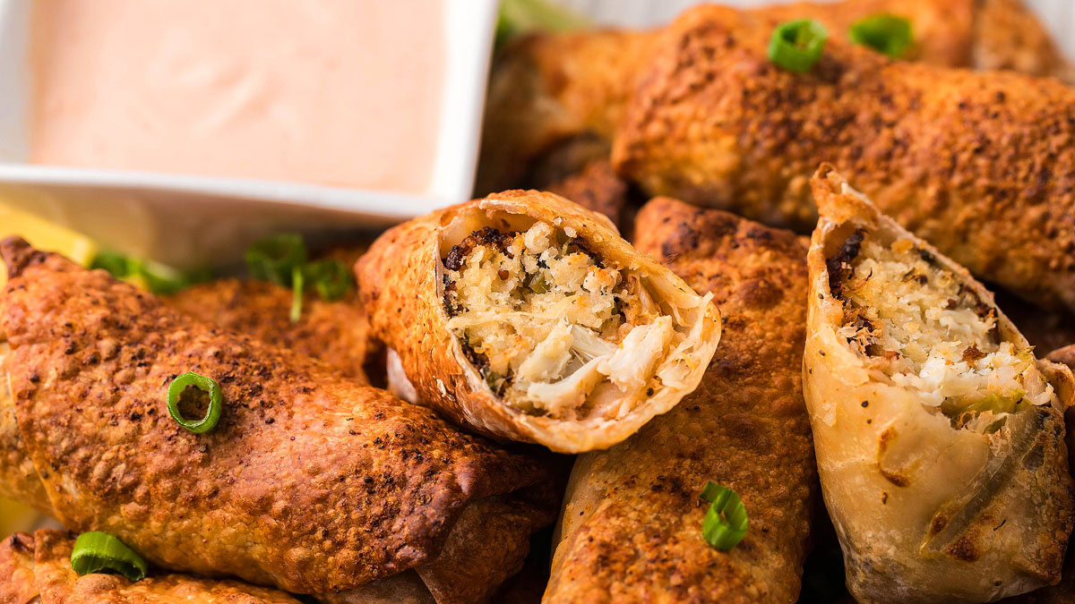 A close-up of crispy egg rolls filled with a shredded mixture, garnished with chopped green onions, next to a dish of creamy dipping sauce.