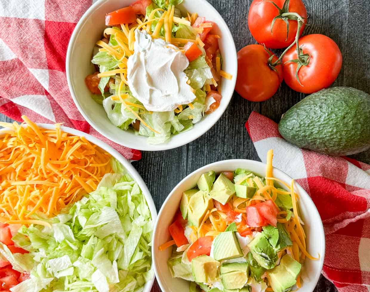 Two bowls of salad with lettuce, tomato, avocado, shredded cheese, and sour cream, next to whole tomatoes, an avocado, and a bowl of lettuce and cheese on a red checkered cloth.