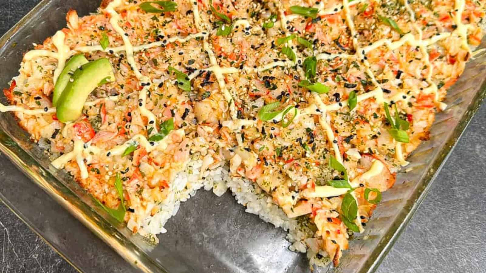 A glass baking dish filled with sushi bake topped with crab, sliced green onions, sesame seeds, and drizzled sauce; a square piece is missing from one corner.