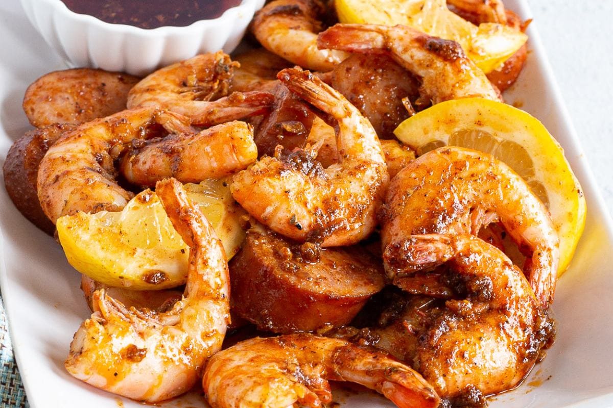 A plate of seasoned shrimp, sausage slices, and lemon wedges, served with a small dish of dipping sauce.