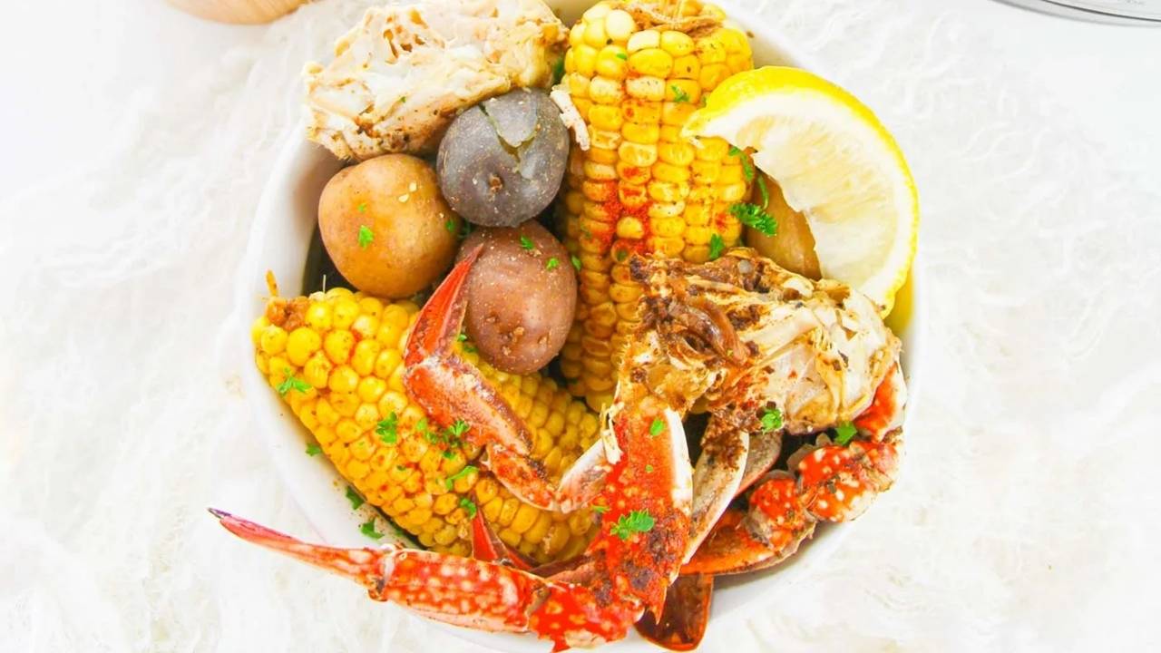 A bowl containing crab claws, corn on the cob, whole potatoes, and a lemon wedge, garnished with parsley.