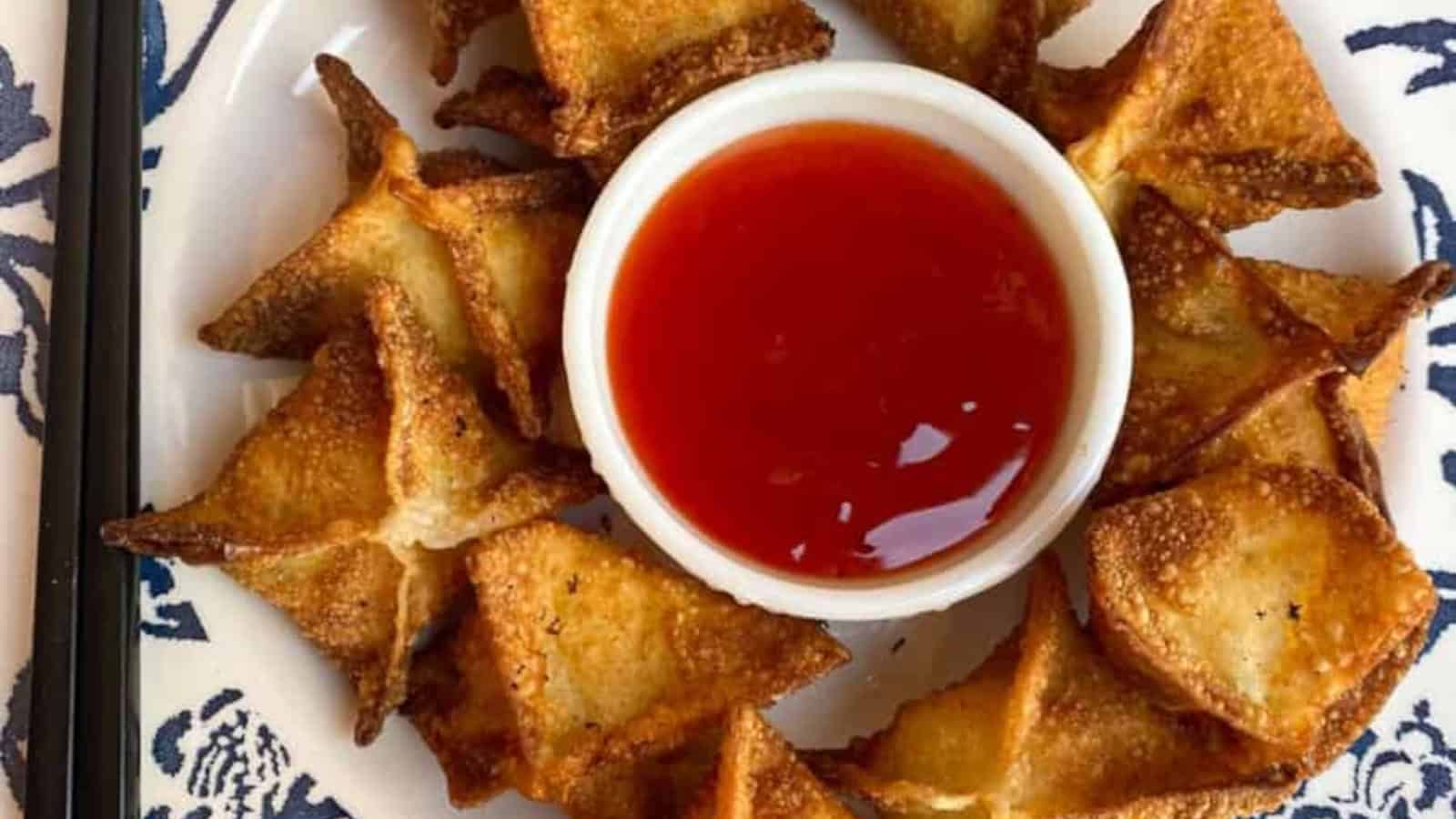 A white plate with crispy fried wontons arranged in a circle around a small bowl of red dipping sauce.