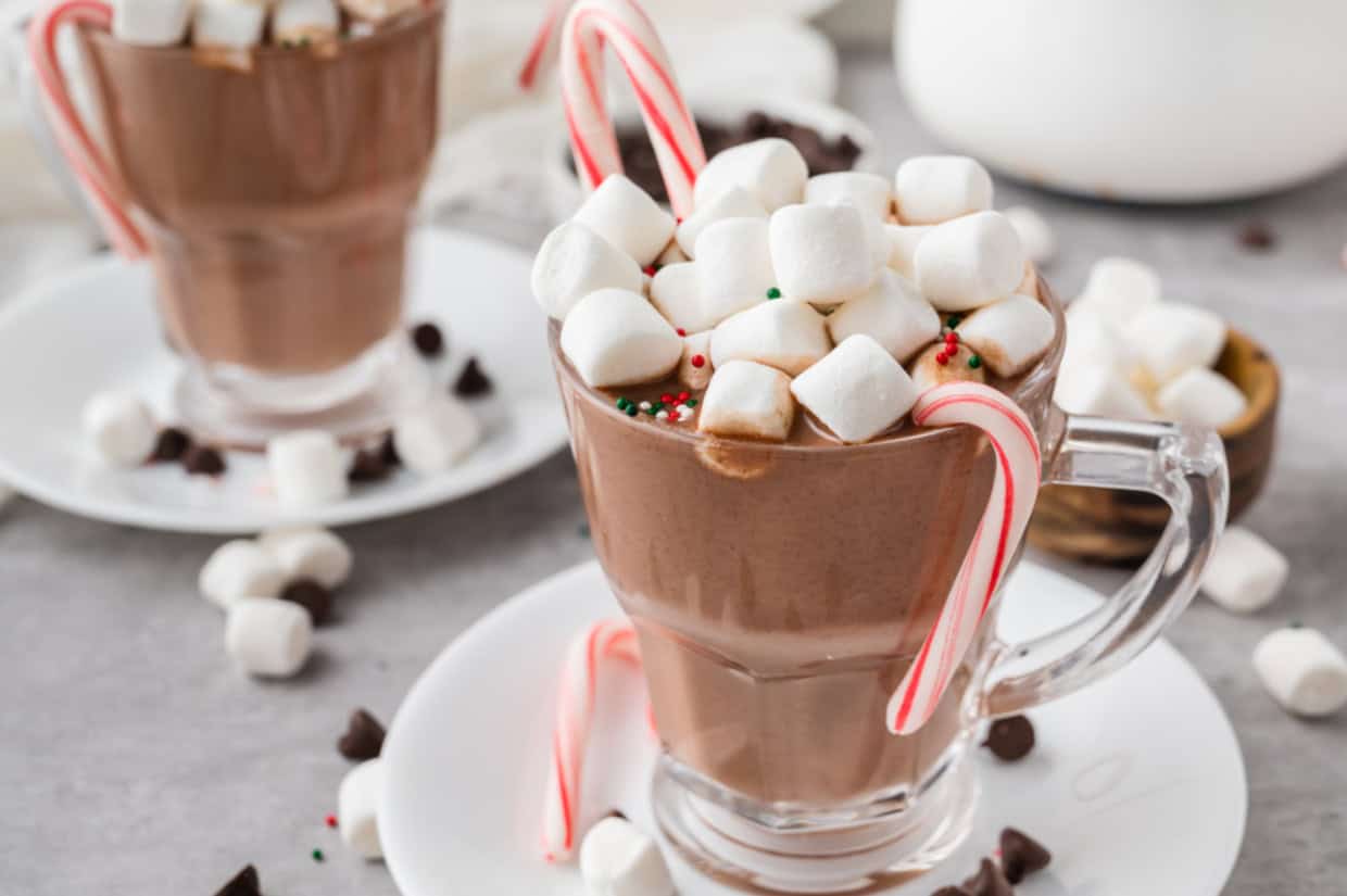 A glass mug of hot chocolate topped with mini marshmallows, candy cane, and sprinkles sits on a white saucer, with another mug in the background.