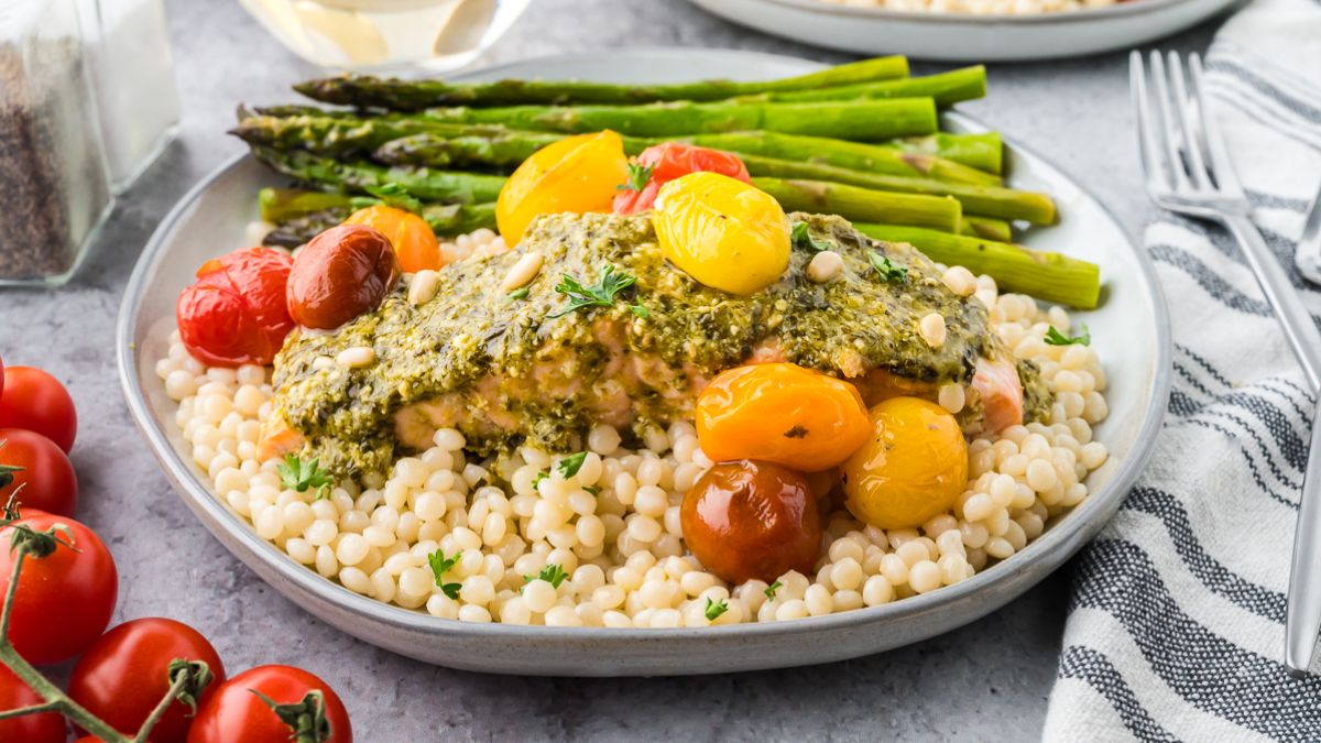 Plate with couscous, pesto-topped fish, roasted cherry tomatoes, and asparagus spears, served with a fork and striped napkin.