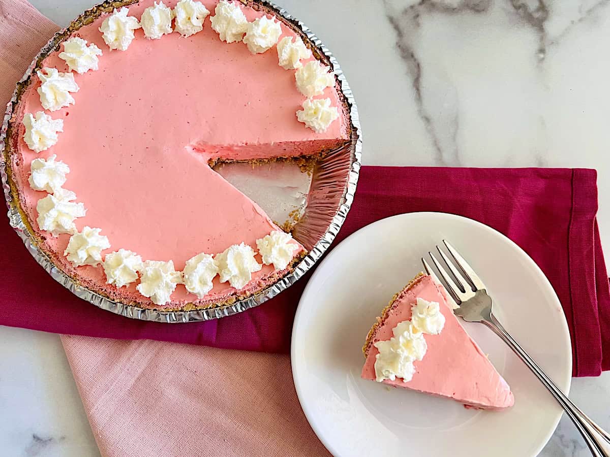 A pink cream pie in a foil pan with a slice missing, topped with whipped cream, sits next to a plate holding the slice and a fork on a pink napkin.