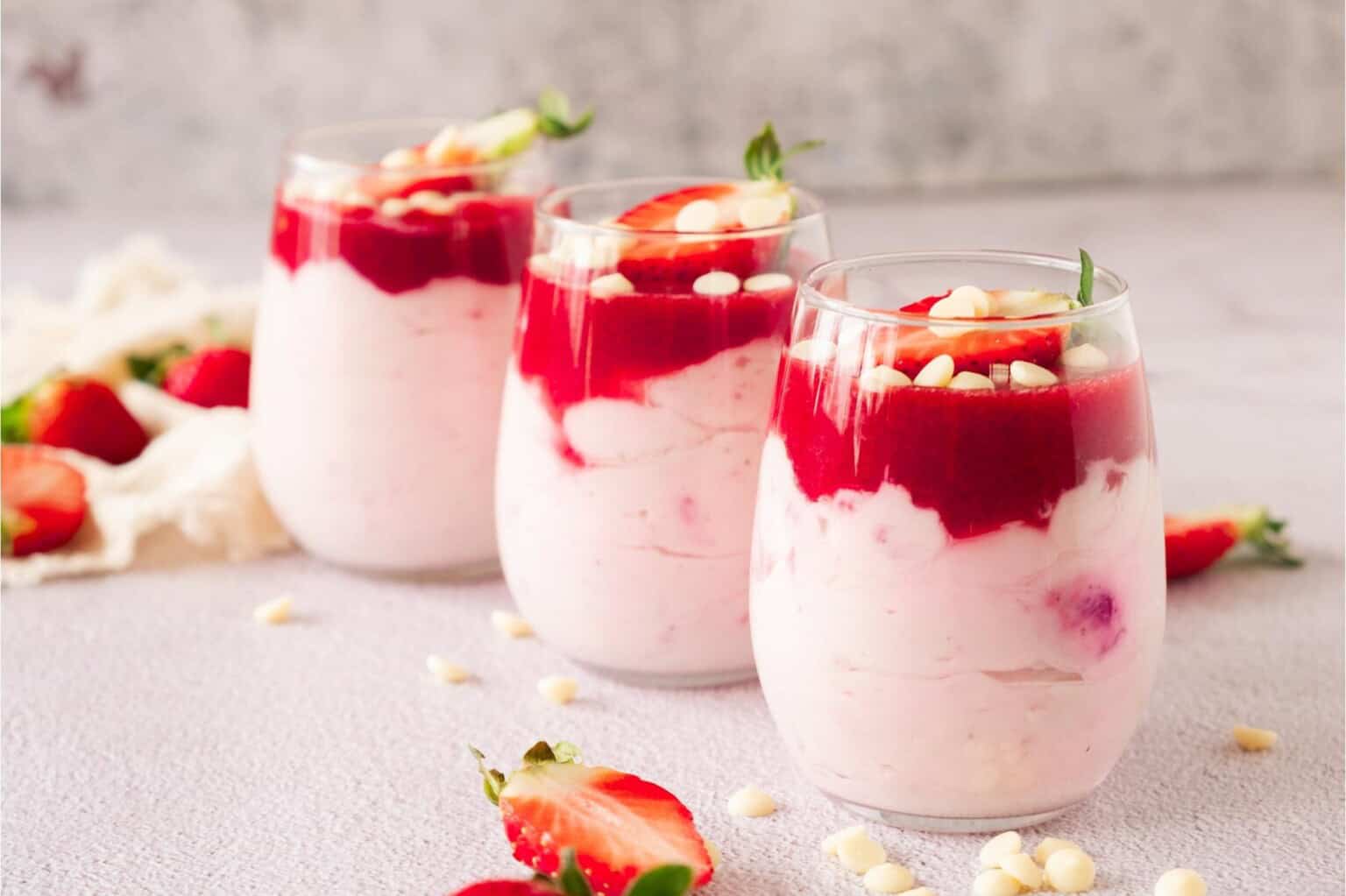 Three glasses of strawberry parfait topped with strawberry sauce, white chocolate chips, and strawberry slices, arranged on a light surface.