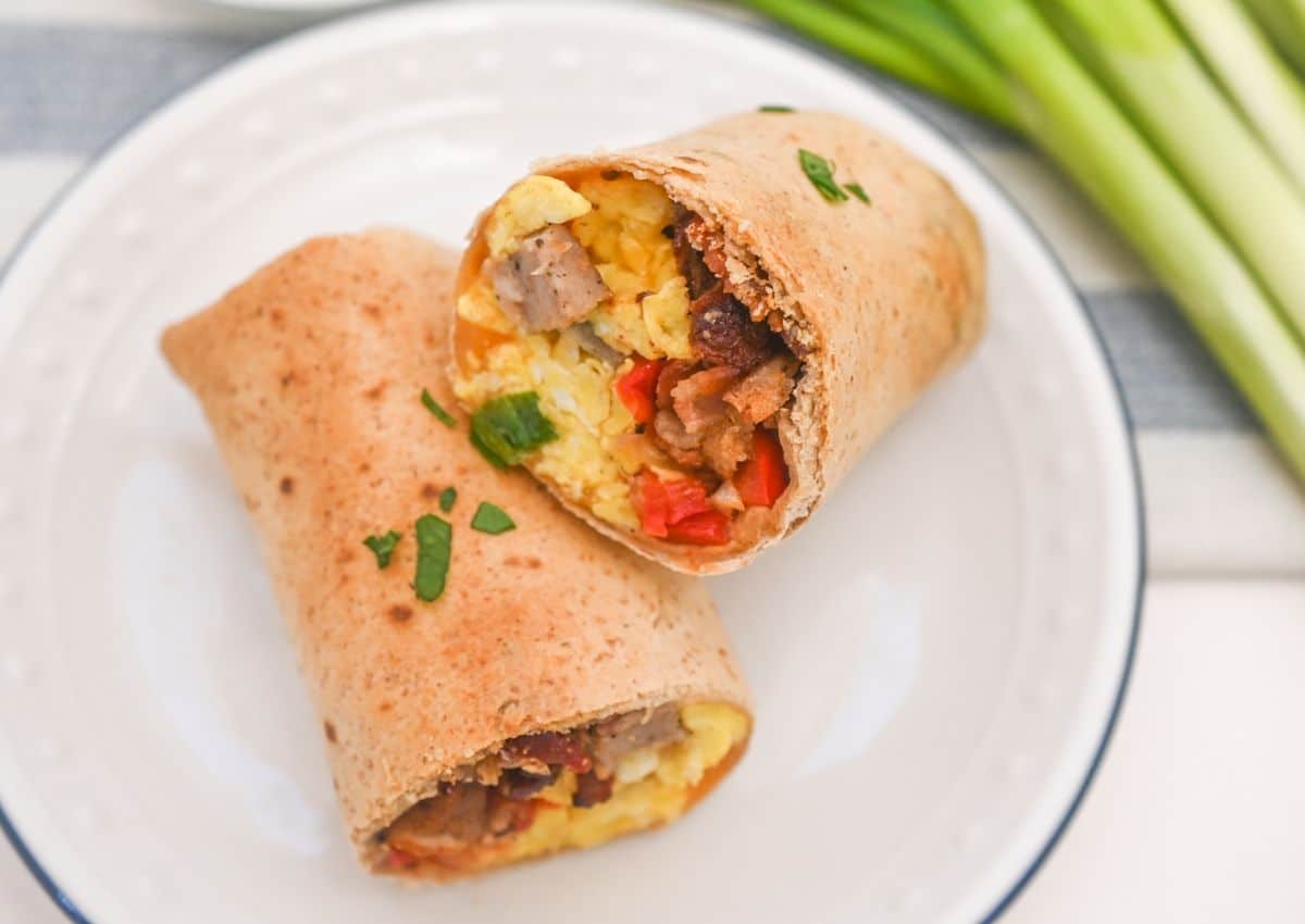 A breakfast burrito cut in half on a white plate, filled with scrambled eggs, sausage, red bell peppers, and garnished with chopped herbs. Green onions are in the background.