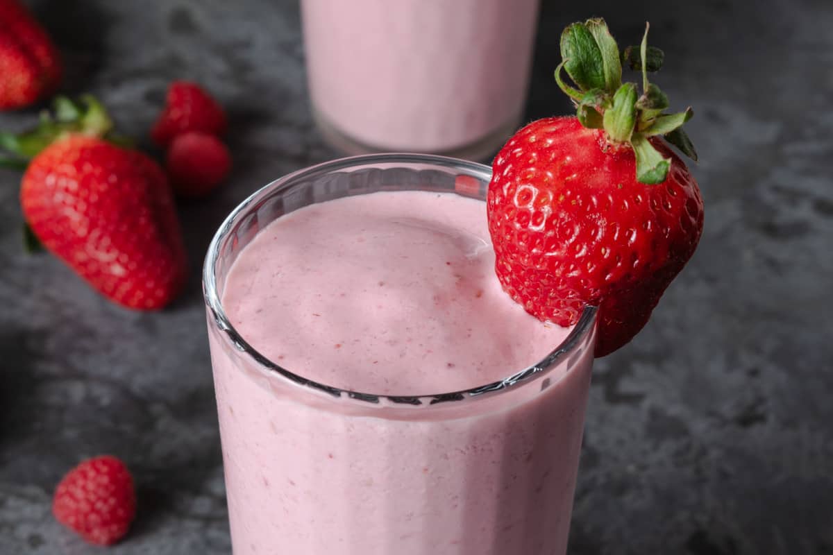 A glass of pink strawberry smoothie topped with a whole strawberry, with fresh strawberries and raspberries in the background on a dark surface.