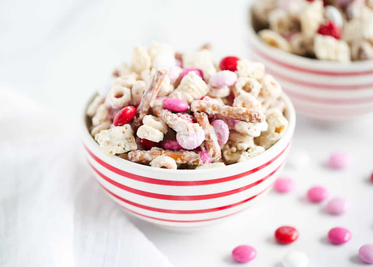 A bowl with red and white stripes filled with a snack mix of pretzels, cereal, and pink, red, and white candy-coated chocolates. Some candies are scattered nearby.