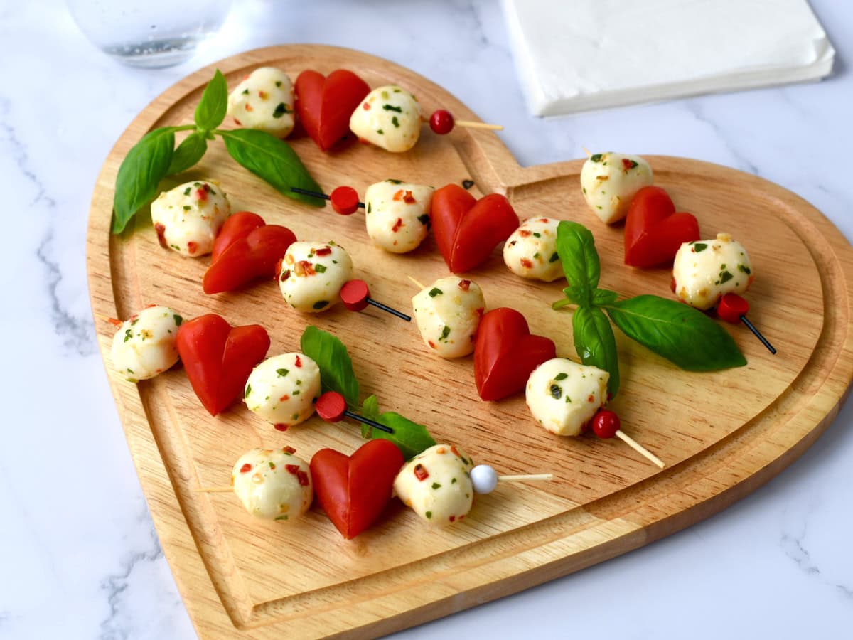 Two skewers with mozzarella balls, heart-shaped cherry tomatoes, and basil leaves are arranged on a heart-shaped wooden board.