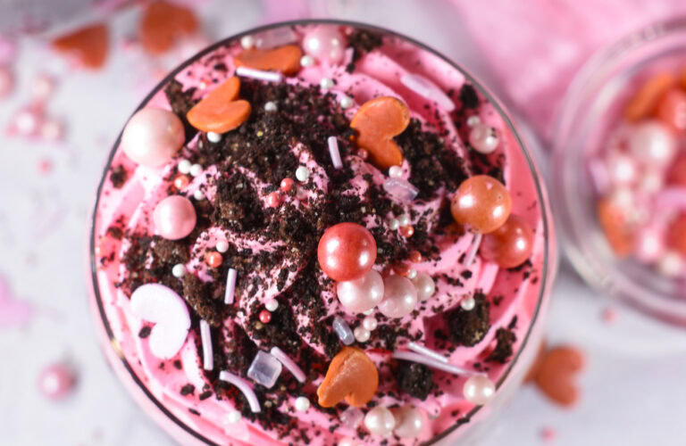 A close-up of a dessert topped with pink whipped cream, chocolate crumbs, pearl-like candies, and heart-shaped sprinkles.