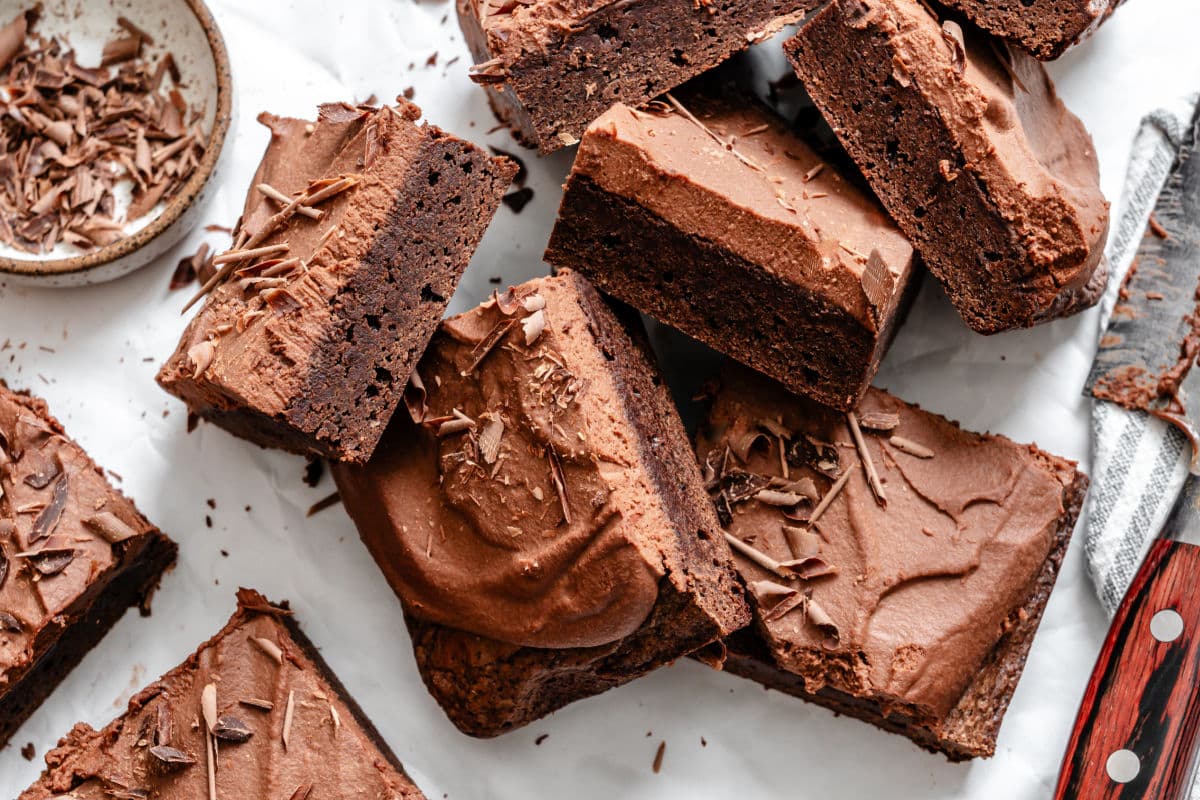 Several frosted chocolate brownies are arranged on a white surface, some topped with chocolate shavings, with a small bowl of shavings and a knife nearby.