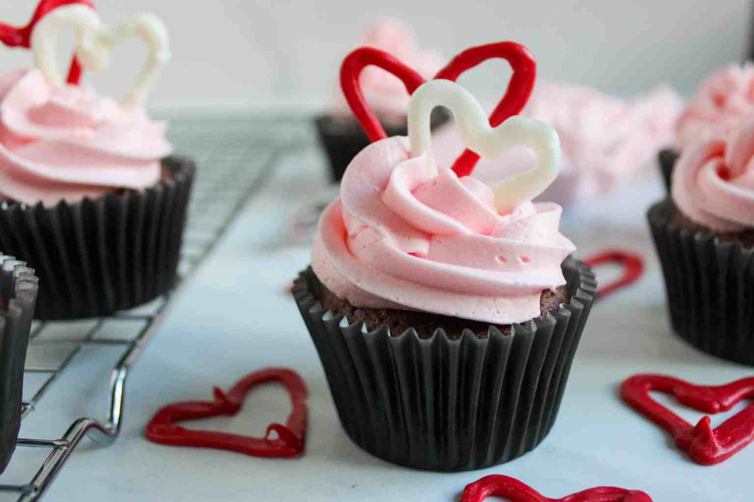 Chocolate cupcakes with pink frosting are topped with red and white chocolate heart decorations, displayed on a cooling rack and a white surface.
