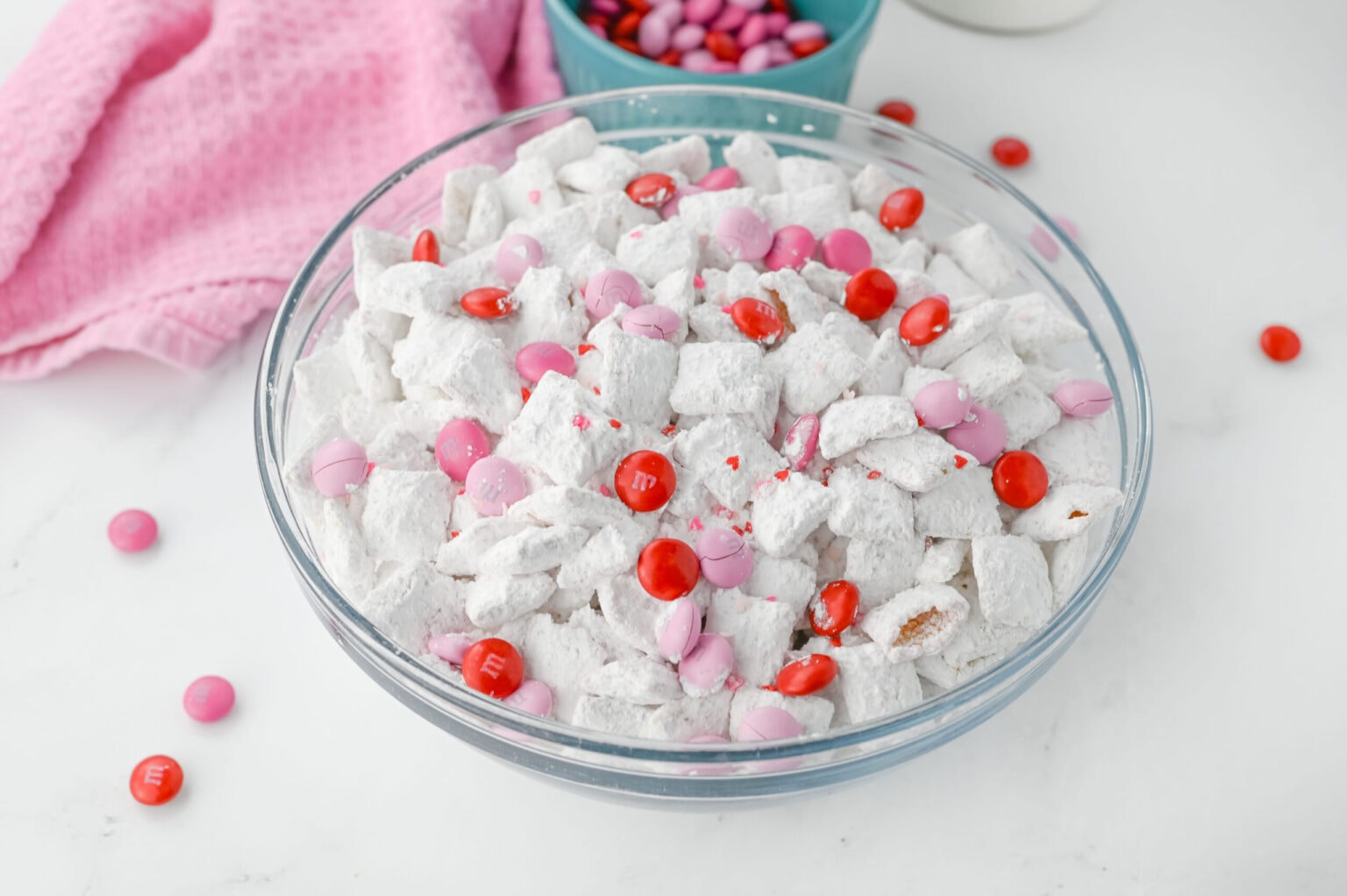 A glass bowl filled with powdered sugar-coated snack mix topped with pink and red candy-coated chocolates, with a pink cloth and candies in the background.