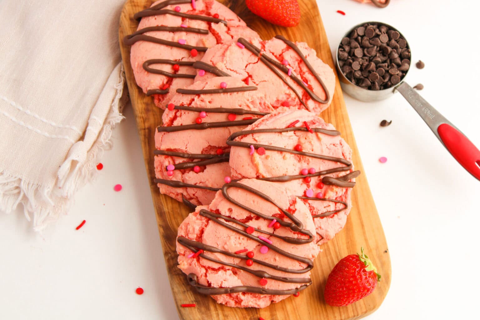 A wooden board with several pink cookies drizzled with chocolate, garnished with heart-shaped sprinkles, surrounded by strawberries and a measuring cup of chocolate chips.