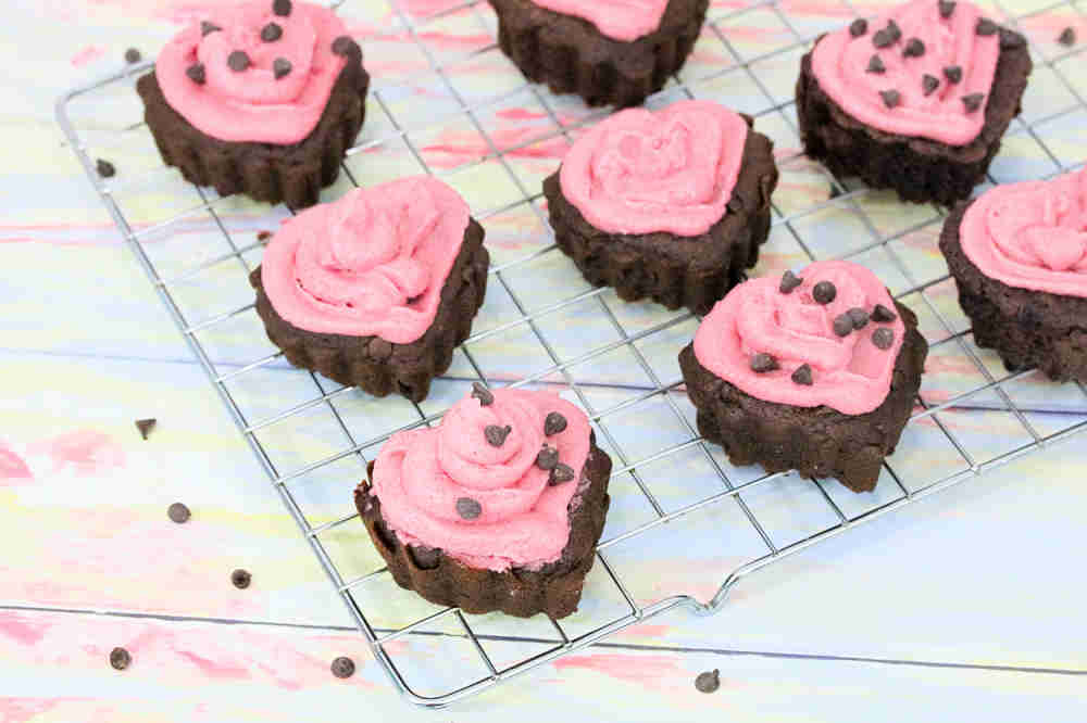 Heart-shaped chocolate brownies with pink frosting and chocolate chips are arranged on a cooling rack.