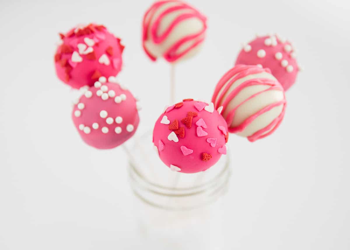 Five cake pops decorated with pink, red, and white icing and sprinkles are arranged in a glass jar against a white background.