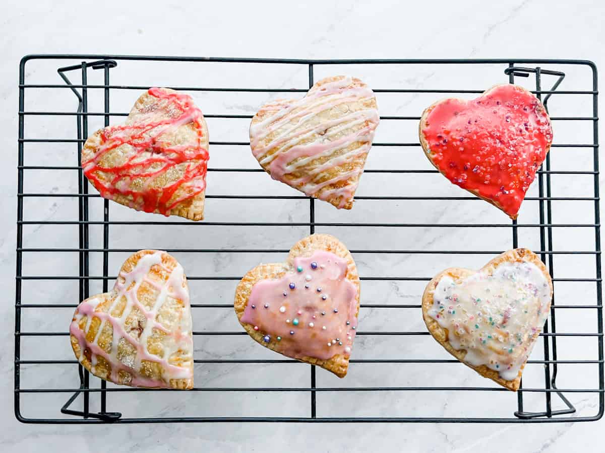 Six heart-shaped pastries with colorful icing and sprinkles are arranged on a black cooling rack over a white surface.
