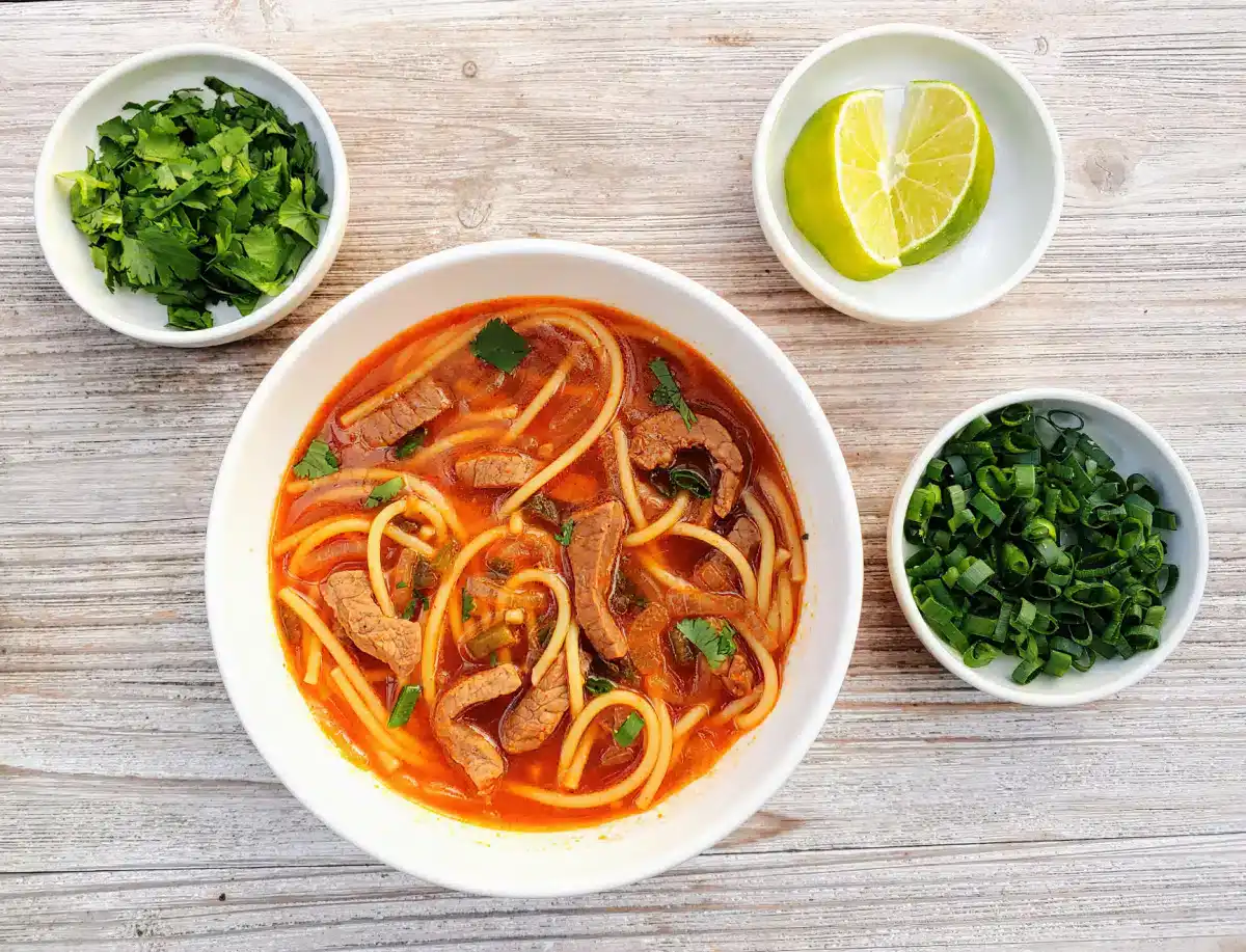 A bowl of beef noodle soup with herbs, accompanied by small bowls of chopped cilantro, sliced green onions, and a lime wedge on a wooden surface.
