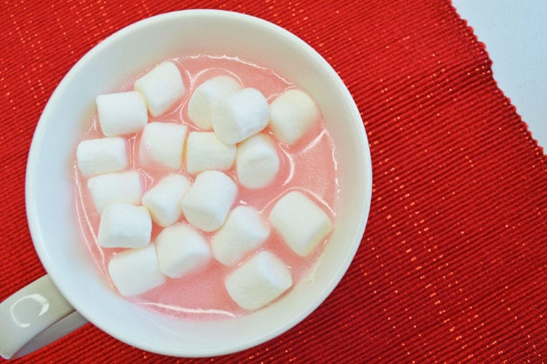 A white mug filled with pink liquid topped with mini marshmallows sits on a red textured placemat.