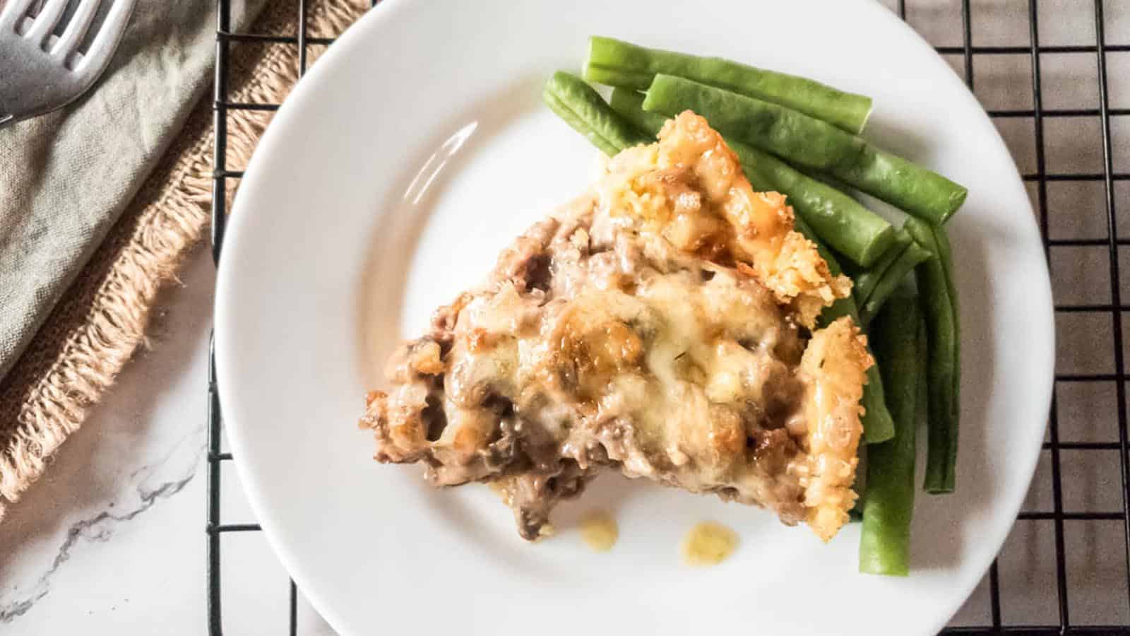 A slice of casserole topped with melted cheese and a side of green beans on a white plate.