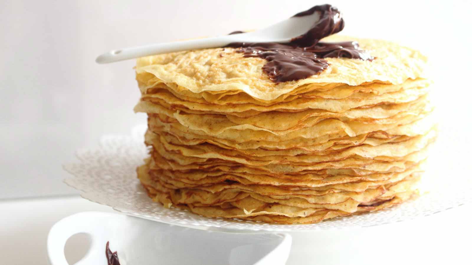 A stack of thin crepes on a white plate with a white spoon and chocolate spread on top.