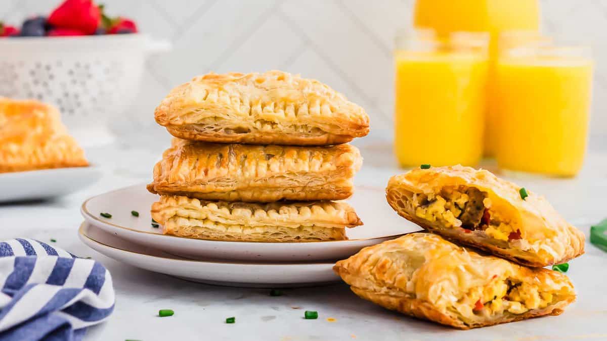 Three puff pastry breakfast pockets stacked on a plate, with two more pockets beside itโone cut open showing scrambled eggs filling. Glasses of orange juice and berries are in the background.