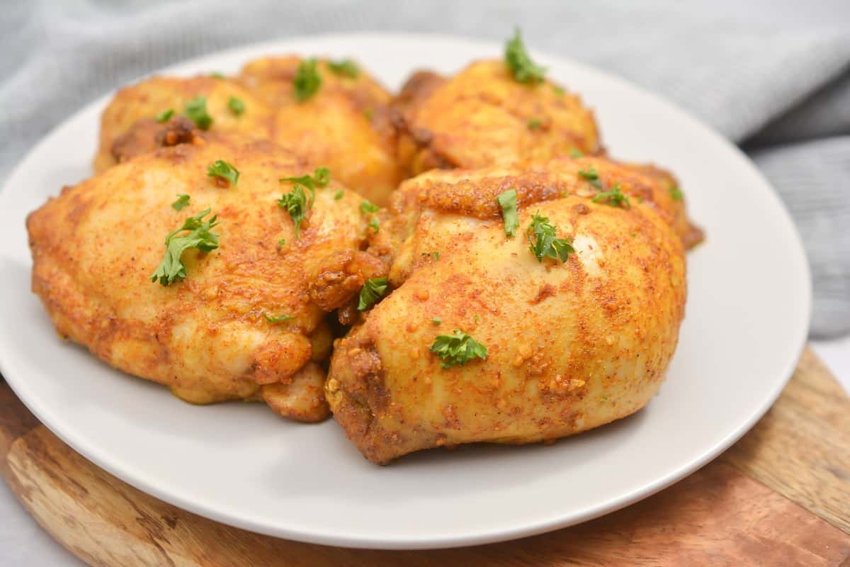 Four seasoned, baked chicken thighs garnished with chopped parsley are arranged on a white plate.