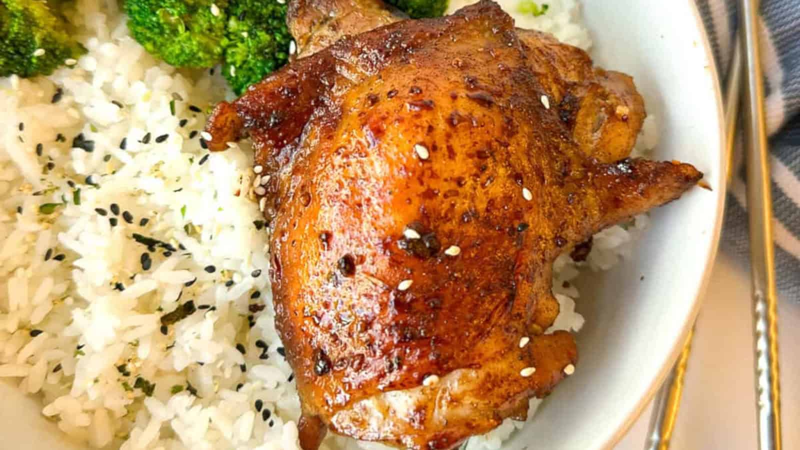 A bowl of white rice topped with a seasoned baked chicken thigh, sprinkled with sesame seeds, and served with broccoli.