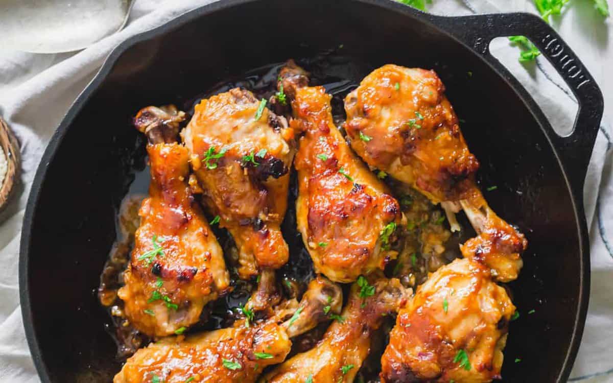 Cooked chicken drumsticks with a golden brown glaze are arranged in a black cast iron skillet, garnished with chopped herbs.
