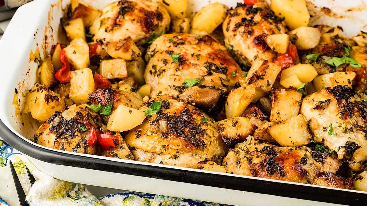 Baked chicken pieces with roasted potatoes, red bell peppers, and herbs in a white baking dish.