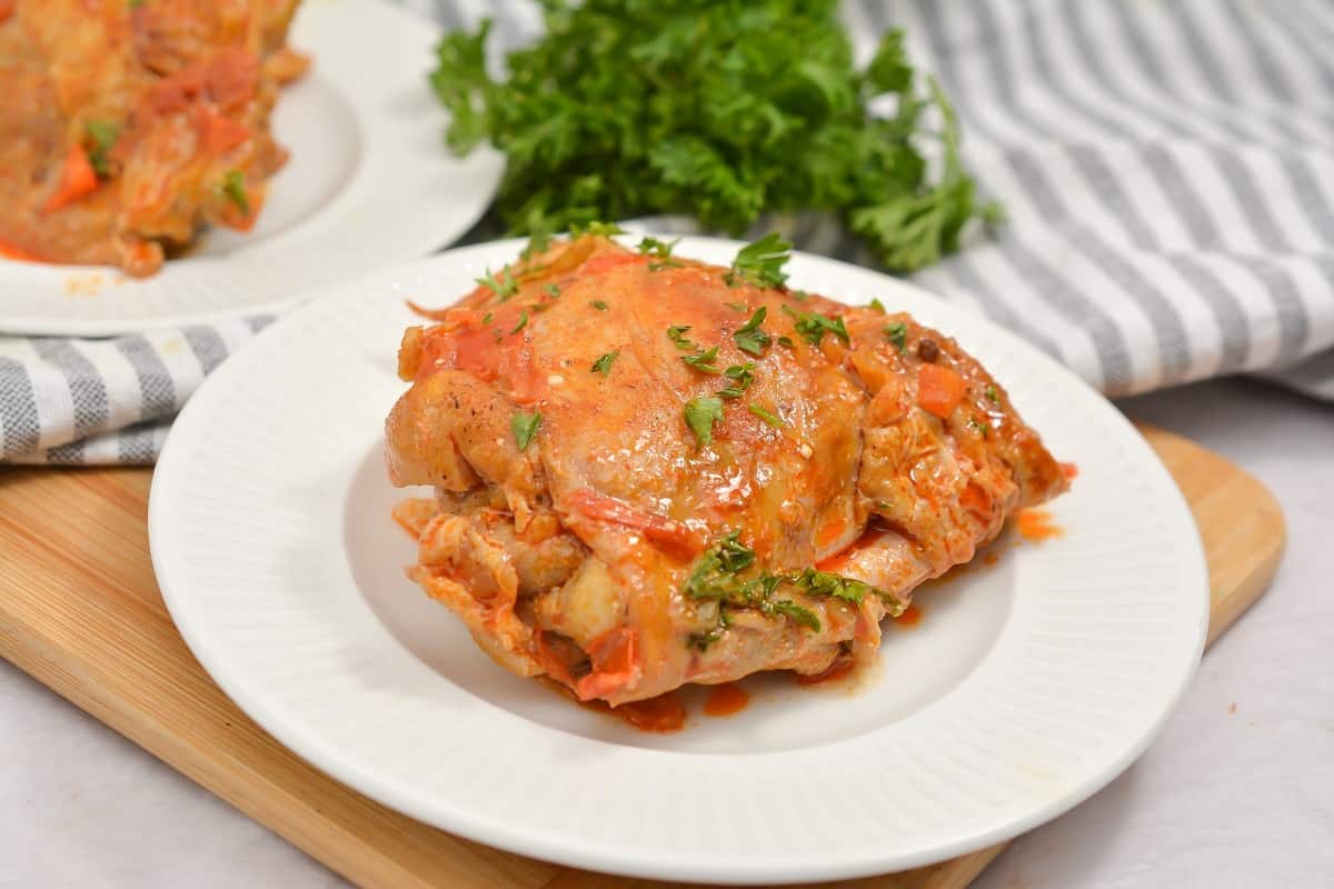 A serving of baked chicken covered in tomato sauce and garnished with chopped herbs on a white plate, with parsley and a striped cloth in the background.