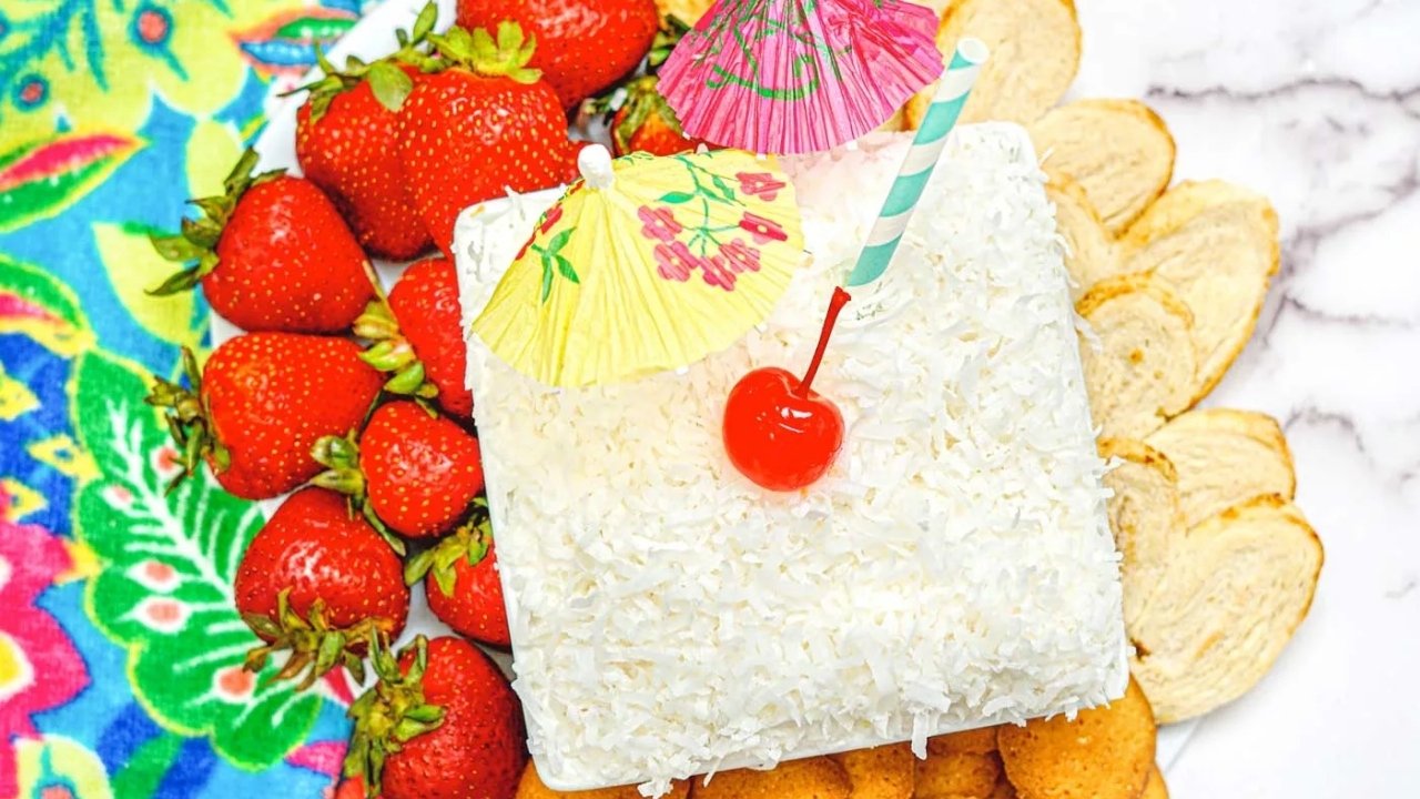 A coconut-covered square dessert with a cherry, paper umbrellas, and straw is surrounded by strawberries, cookies, and sliced bread on a floral-patterned cloth.