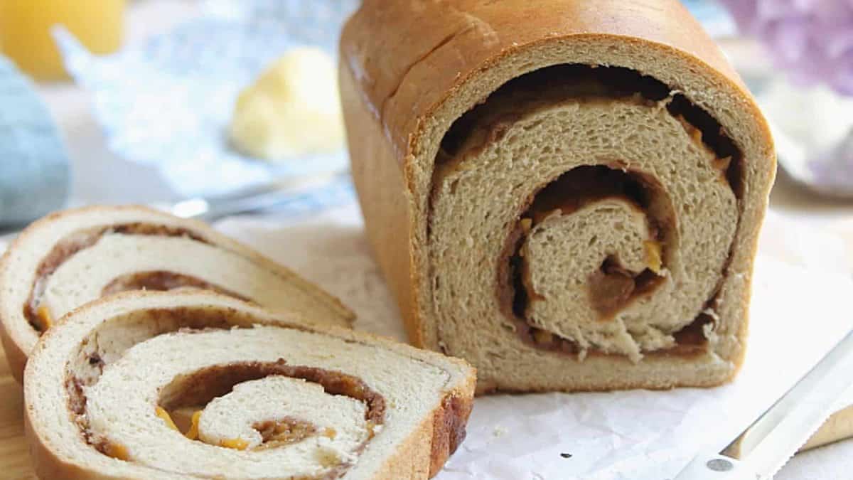 A loaf of cinnamon swirl bread with two slices cut, showing the spiral pattern inside. A butter knife is placed nearby on a parchment-covered surface.
