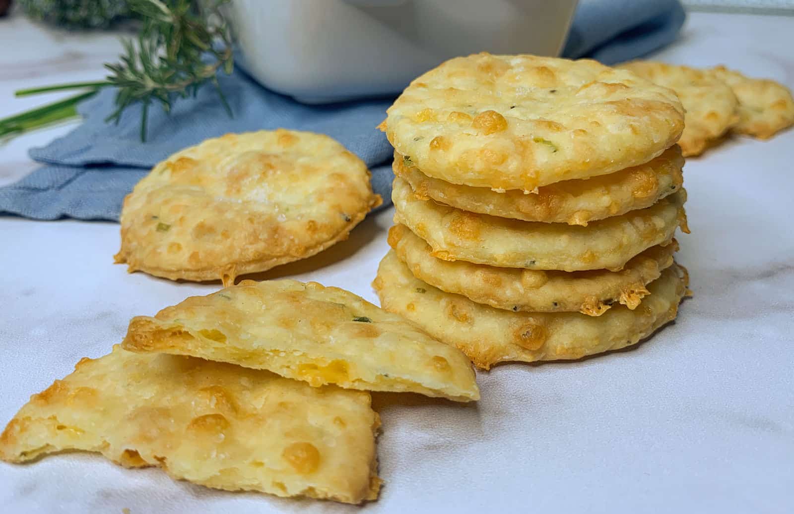 A stack of round, golden cheese crackers is shown on a white surface, with a few broken pieces and a blue cloth in the background.