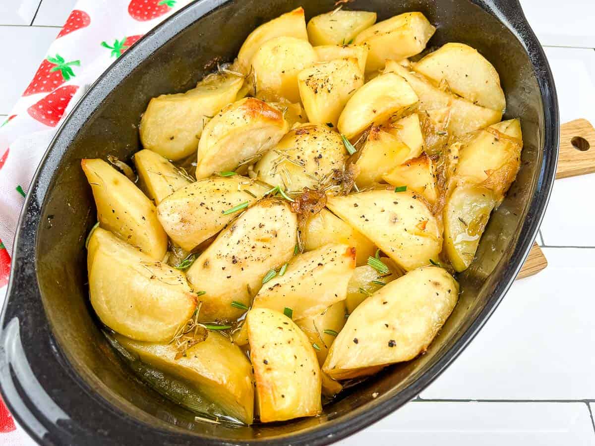 Sliced roasted potatoes seasoned with herbs and black pepper, served in a black baking dish on a white tiled surface.