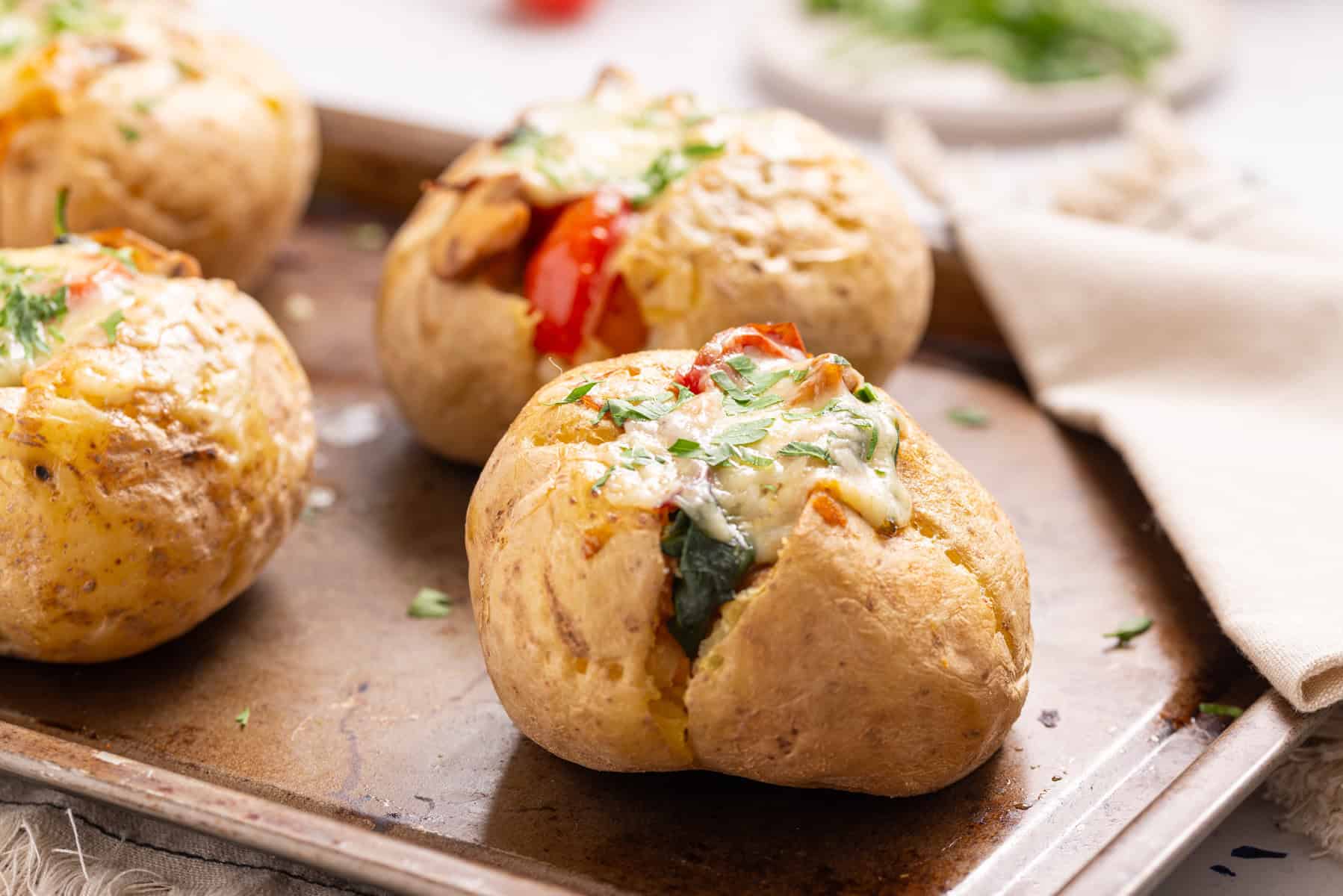 Several baked potatoes topped with melted cheese, vegetables, and herbs are arranged on a baking tray.