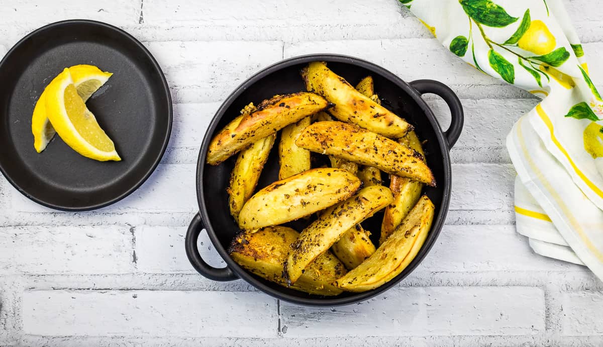 A black bowl filled with seasoned potato wedges sits on a white brick surface next to a small plate with two lemon wedges and a lemon-patterned cloth.